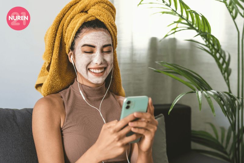 girl using mask on her face with towel turban dring her hair and listening to music on her earphones connected to the iphone