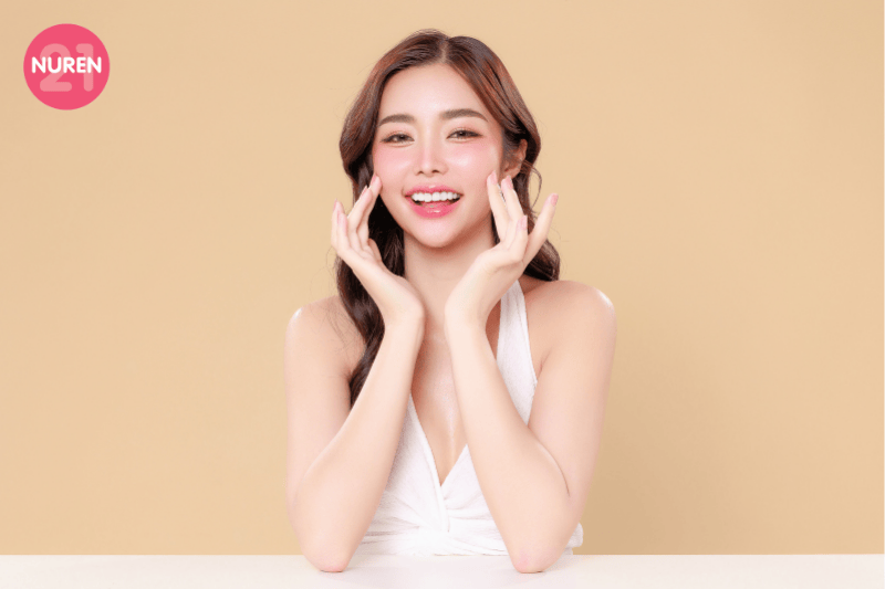 woman holding her face in front of a beige screen looking into the camera and smiling looking happy