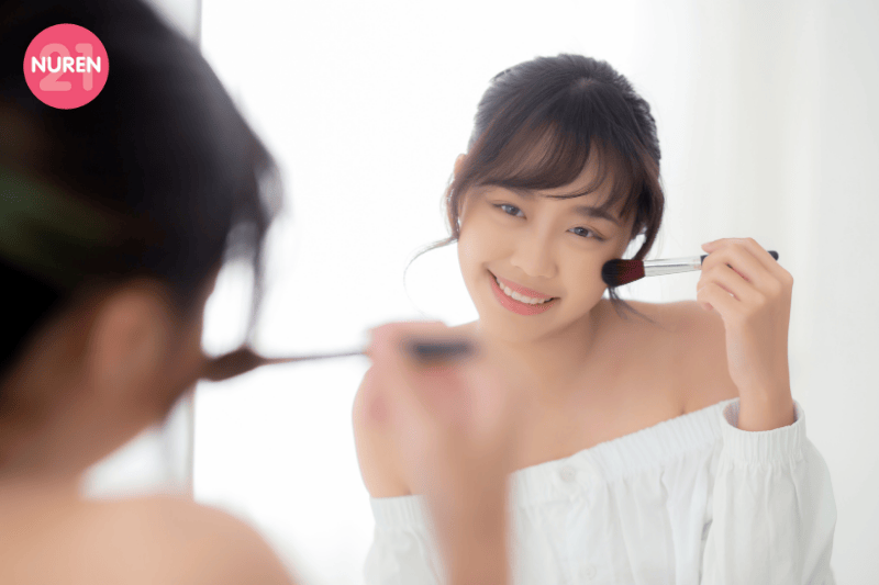 image of young woman putting light blush on face and looking at the mirror