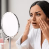 Shocked young indian woman looking at her face at mirror, wearing white bathrobe, sitting next to window in bathroom, touching her skin, suffering from puffy eyes in the morning