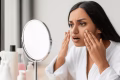 Shocked young indian woman looking at her face at mirror, wearing white bathrobe, sitting next to window in bathroom, touching her skin, suffering from puffy eyes in the morning