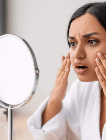 Shocked young indian woman looking at her face at mirror, wearing white bathrobe, sitting next to window in bathroom, touching her skin, suffering from puffy eyes in the morning