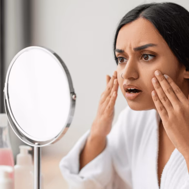 Shocked young indian woman looking at her face at mirror, wearing white bathrobe, sitting next to window in bathroom, touching her skin, suffering from puffy eyes in the morning