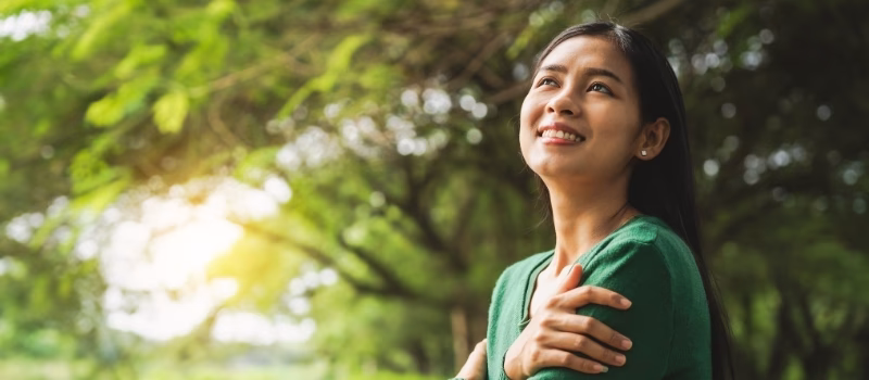 confident asian woman embraces self-love, standing peacefully in serene park at sunset. joyful smile, she enjoys fresh air, breathing deeply, radiates calm and positivity in tranquil surroundings.