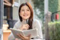 Pretty asian woman holding book thinking with pen under chin sitting at wooden table outside a cafe. Leisure Time, Everyday Life, Chilling Outdoors.