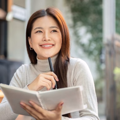 Pretty asian woman holding book thinking with pen under chin sitting at wooden table outside a cafe. Leisure Time, Everyday Life, Chilling Outdoors.