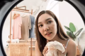 Young Asian woman holds white knitwear near washing machine door, friendly home lifestyle laundry preparation, tidy apartment clothing care, weekend domestic chore and organized wardrobe routine