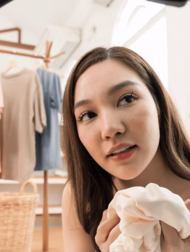 Young Asian woman holds white knitwear near washing machine door, friendly home lifestyle laundry preparation, tidy apartment clothing care, weekend domestic chore and organized wardrobe routine