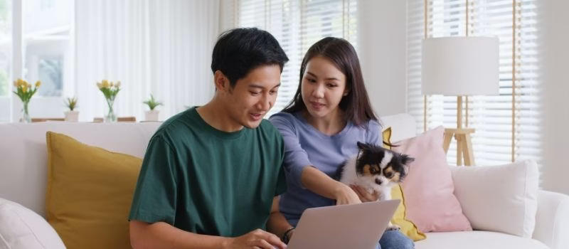 Asia people young family man woman smile happy sit at home sofa couch pay for hotel booking, search choose, buy ticket on laptop social media app travel trip getaway plan tour with cute small pet dog.
