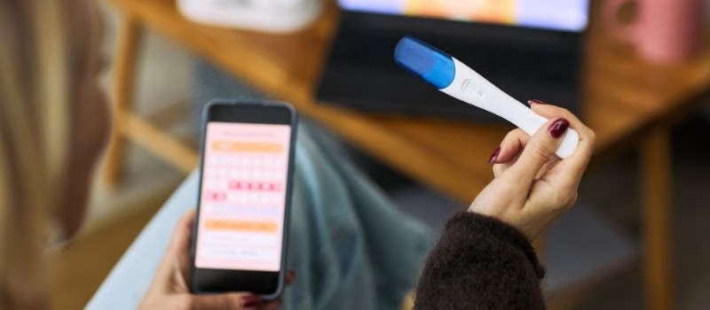 woman holding digital ovulation test and smartphone displaying menstrual cycle tracking app, sitting indoors with laptop in background, focusing on femtech health monitoring