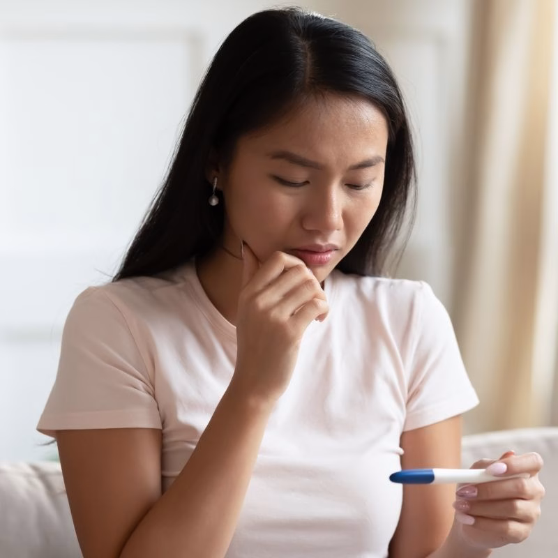 Head shot of asian ethnicity young woman sitting on couch alone in living room feels shocked by positive unplanned with two lines pregnancy test result, abortion decision unwilling pregnancy concept