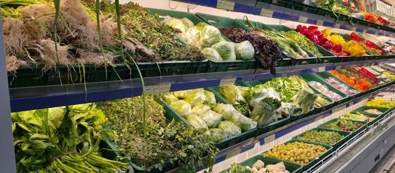Vibrant and well-organized display of fresh vegetables in a modern supermarket. The scene features green leafy vegetables such as spinach, lettuce, and herbs along with colorful bell peppers and root vegetables arranged in plastic crates. Bright indoor lighting enhances the freshness and colors, creating an inviting atmosphere for healthy food shopping and grocery concepts.