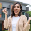 Woman buying or rent new home she holding key front of new house. Surprise happy young asian woman giving house key and smile to rent or purchase apartment home. Moving relocation concept.