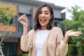 Woman buying or rent new home she holding key front of new house. Surprise happy young asian woman giving house key and smile to rent or purchase apartment home. Moving relocation concept.