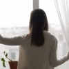 Rear view woman stand near window in living room open curtains looking outside at rainy foggy weather