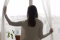 Rear view woman stand near window in living room open curtains looking outside at rainy foggy weather