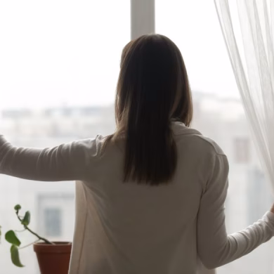 Rear view woman stand near window in living room open curtains looking outside at rainy foggy weather